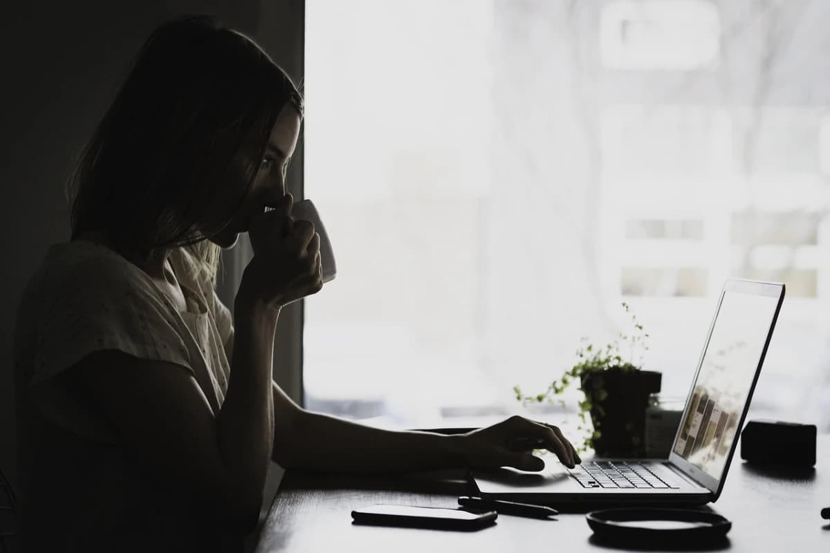 A woman drinking coffee while reading on a laptop by a window.