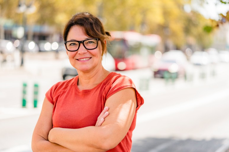Confident middle-aged woman standing outdoors with arms crossed, wearing glasses and a rust-colored t-shirt — representing self-assurance and transformation following weight loss surgery.