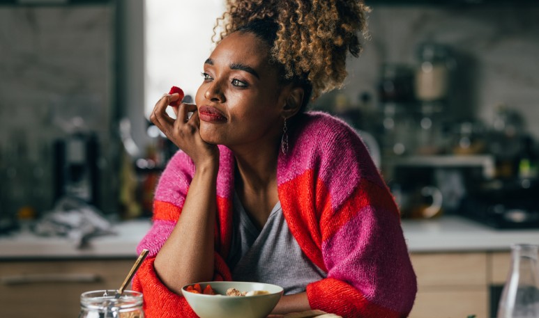 Thoughtful woman sitting at a kitchen counter with a bowl of healthy food, holding a berry near her lips — symbolizing mindful eating and emotional awareness after gastric sleeve surgery.